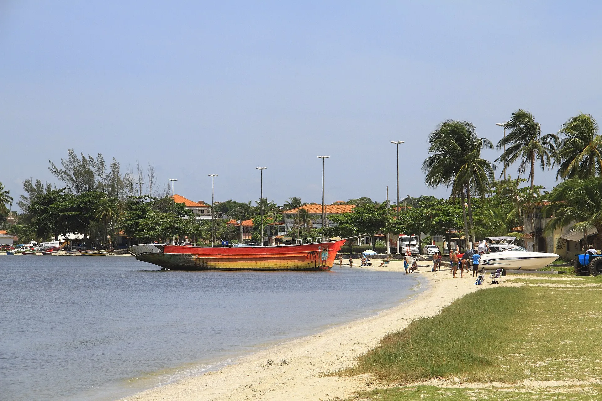Barco de pesca vermelho encostado na areia da orla da lagoa em Iguaba Grande, com coqueiros e casas ao fundo