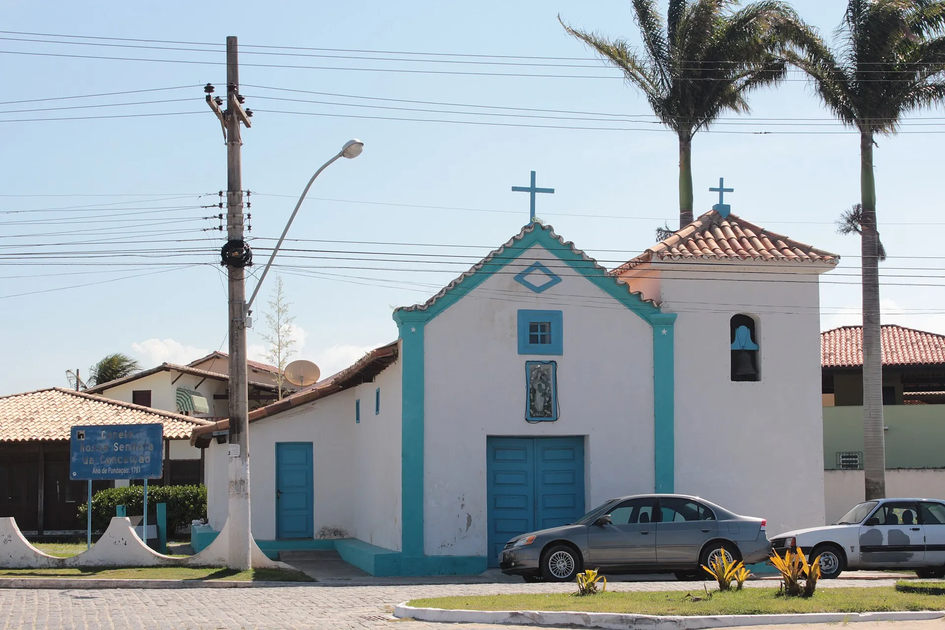 Pequena capela branca de Nossa Senhora da Conceição, com detalhes em azul-turquesa e palmeiras ao fundo, em Iguaba Grande