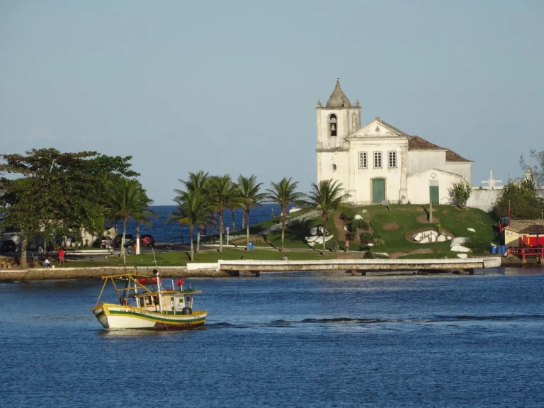 Capela branca de São João Batista no alto de um rochedo à beira-mar, com um barco de pesca passando no rio, em Barra de São João