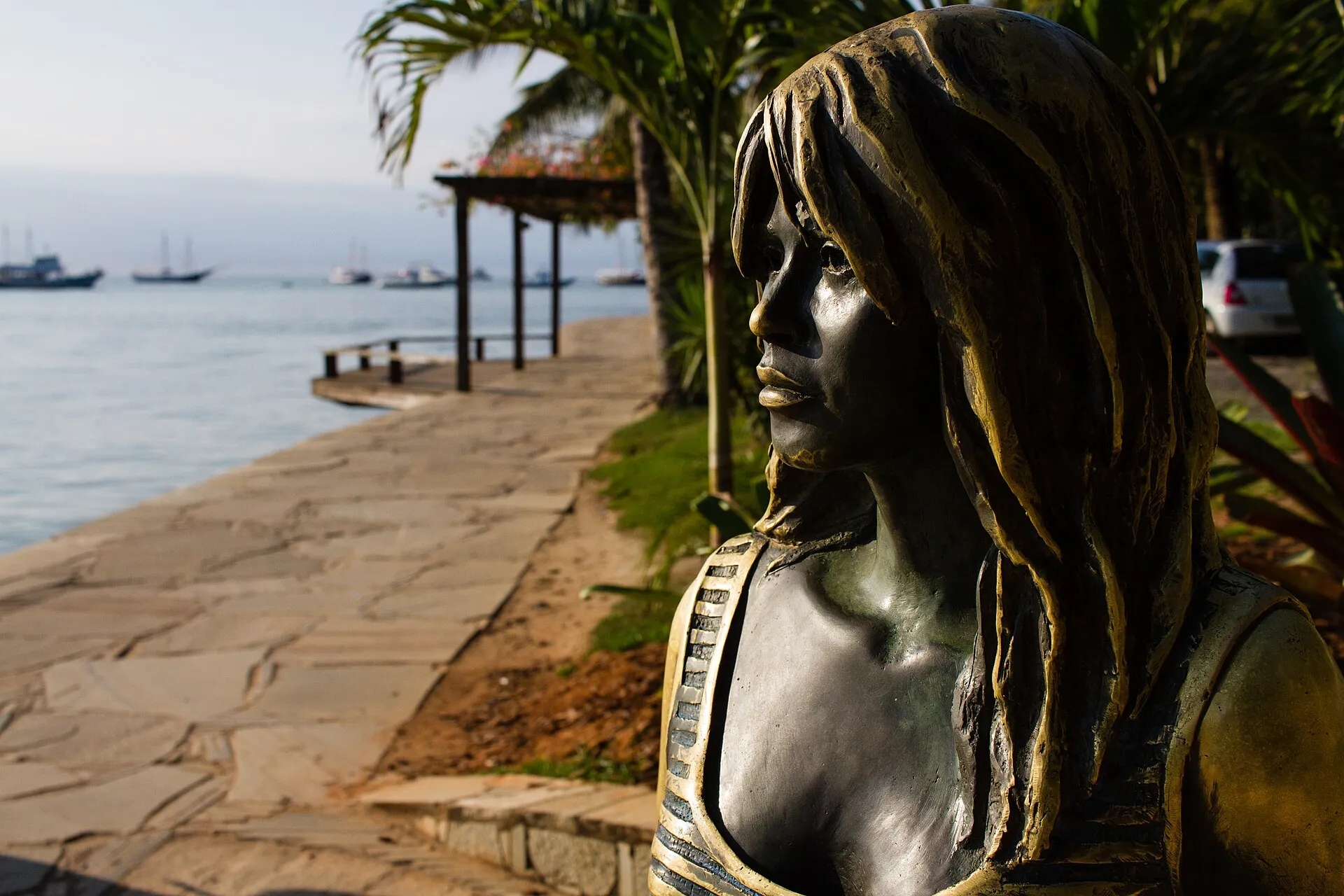 Estátua de bronze de Brigitte Bardot vista de perfil, com o calçadão da Orla Bardot e o mar de Búzios ao fundo