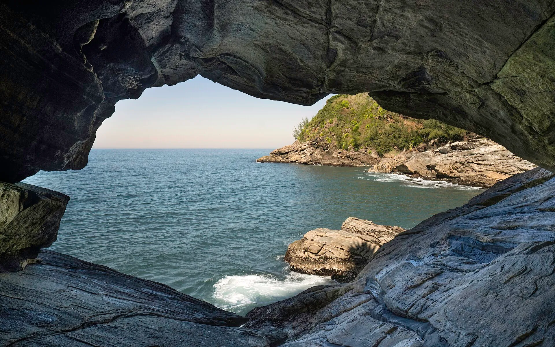 Vista de dentro da Gruta da Sacristia, uma abertura nas rochas emoldurando o mar azul e o costão de Ponta Negra