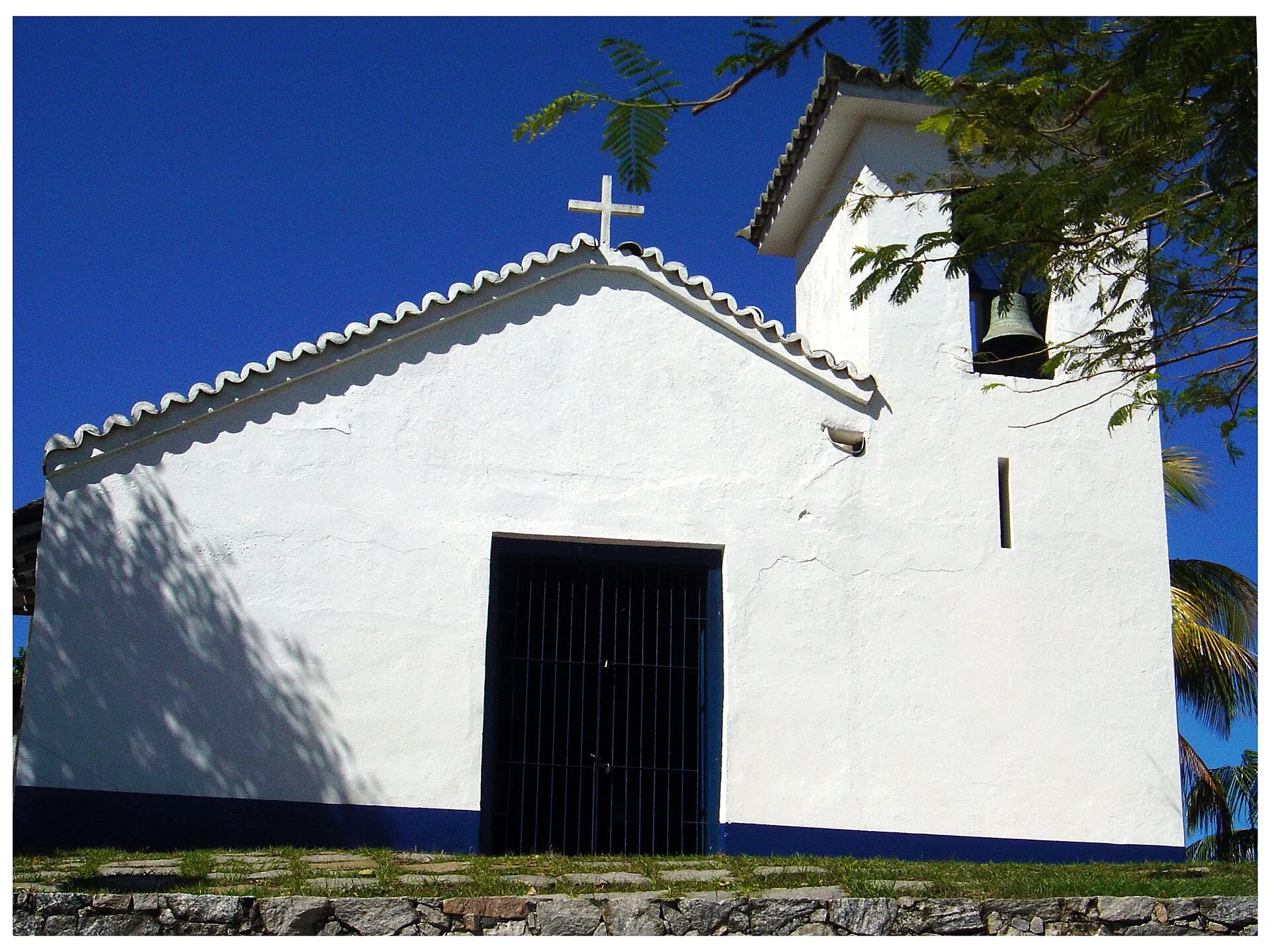 Fachada branca da pequena Igreja de Sant’Anna, com porta escura e campanário lateral, no morro entre a Praia dos Ossos e a Praia da Armação