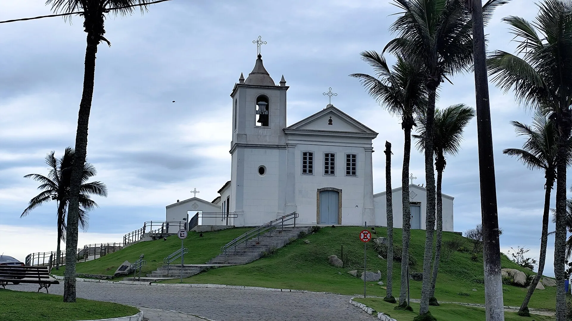 Igreja branca de São João Batista com torre sineira, no alto de um gramado cercado de palmeiras, em Barra de São João