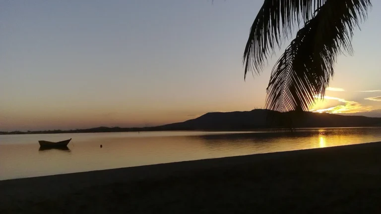 Pôr do sol sobre a Lagoa de Araruama, com uma canoa na água, palmeira em primeiro plano e a Serra de Sapiatiba ao fundo, em São Pedro da Aldeia