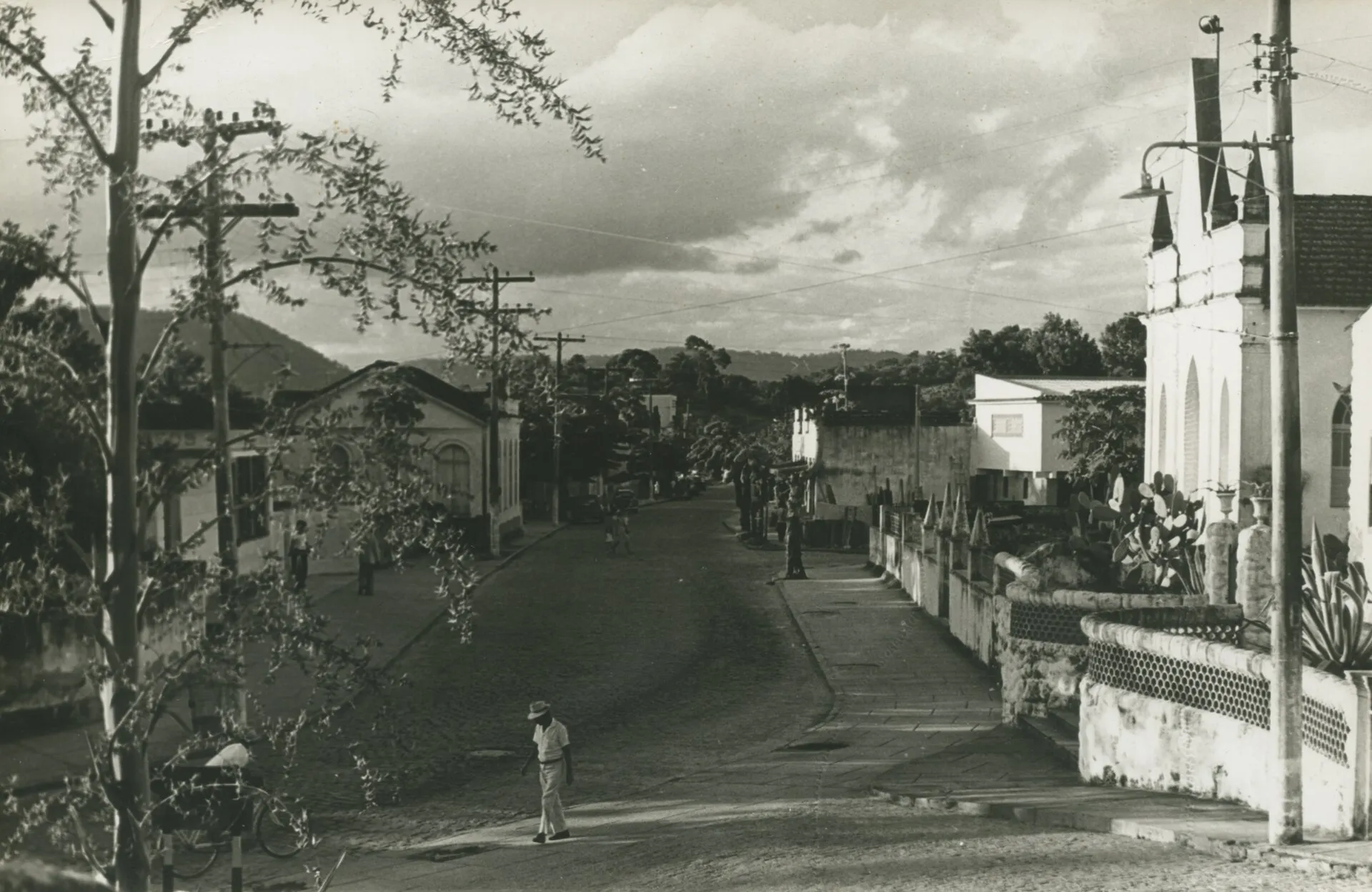 Fotografia em preto e branco de uma rua de Maricá em 1970, com árvores, calçadas e a lateral de uma igreja à direita
