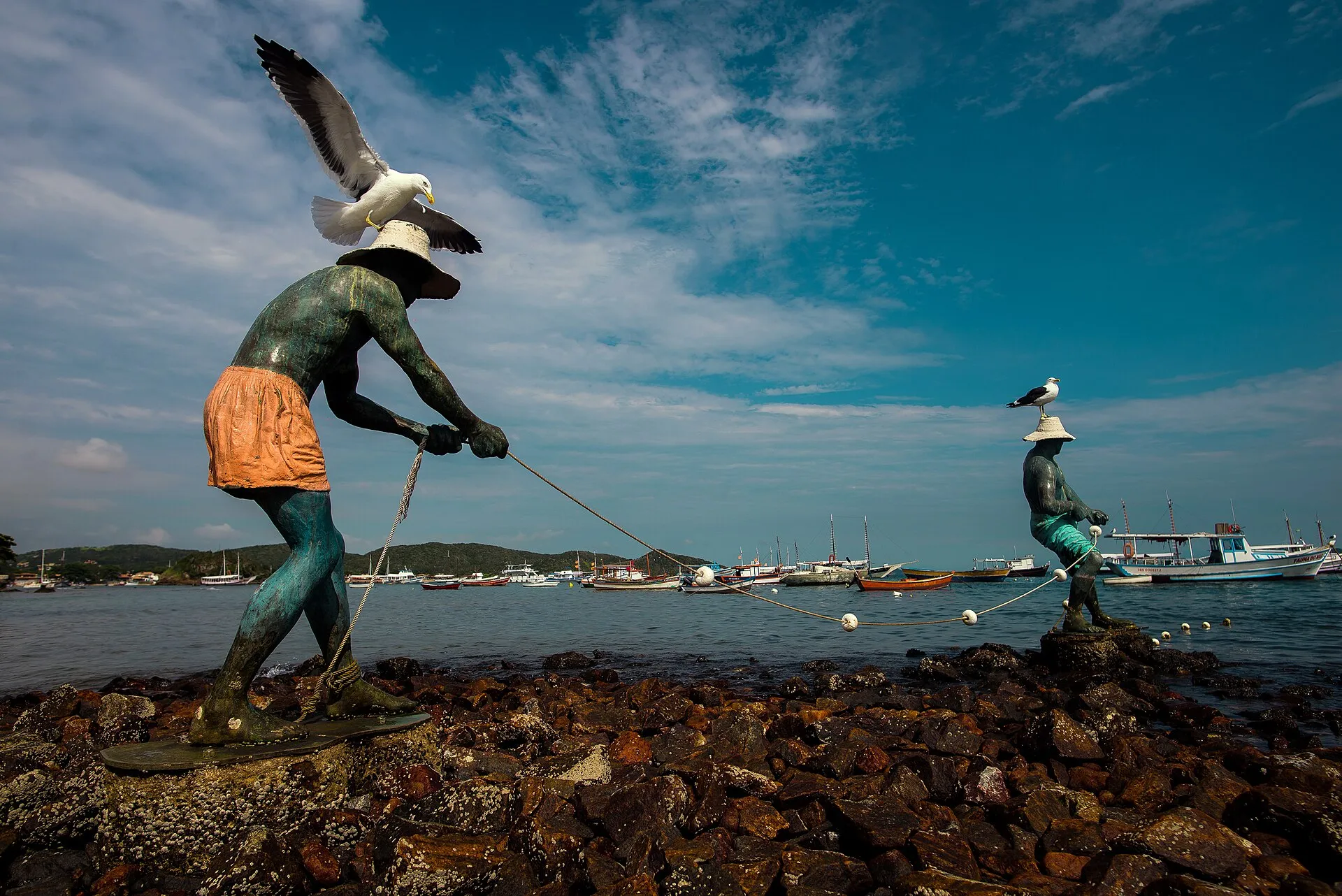 Escultura de um pescador puxando uma corda sobre as pedras à beira-mar, com outra figura do conjunto ao fundo, em Búzios
