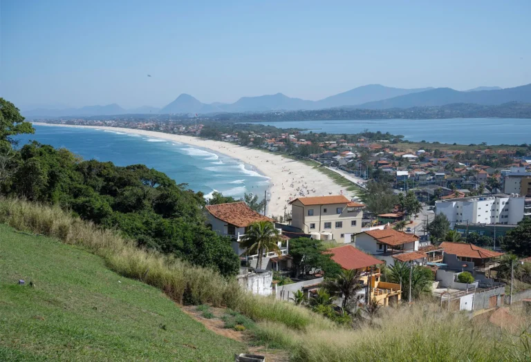 Vista panorâmica do alto do Farol de Ponta Negra, com a longa praia de areia clara, o bairro à beira-mar, a Lagoa de Guarapina e montanhas ao fundo, em Maricá