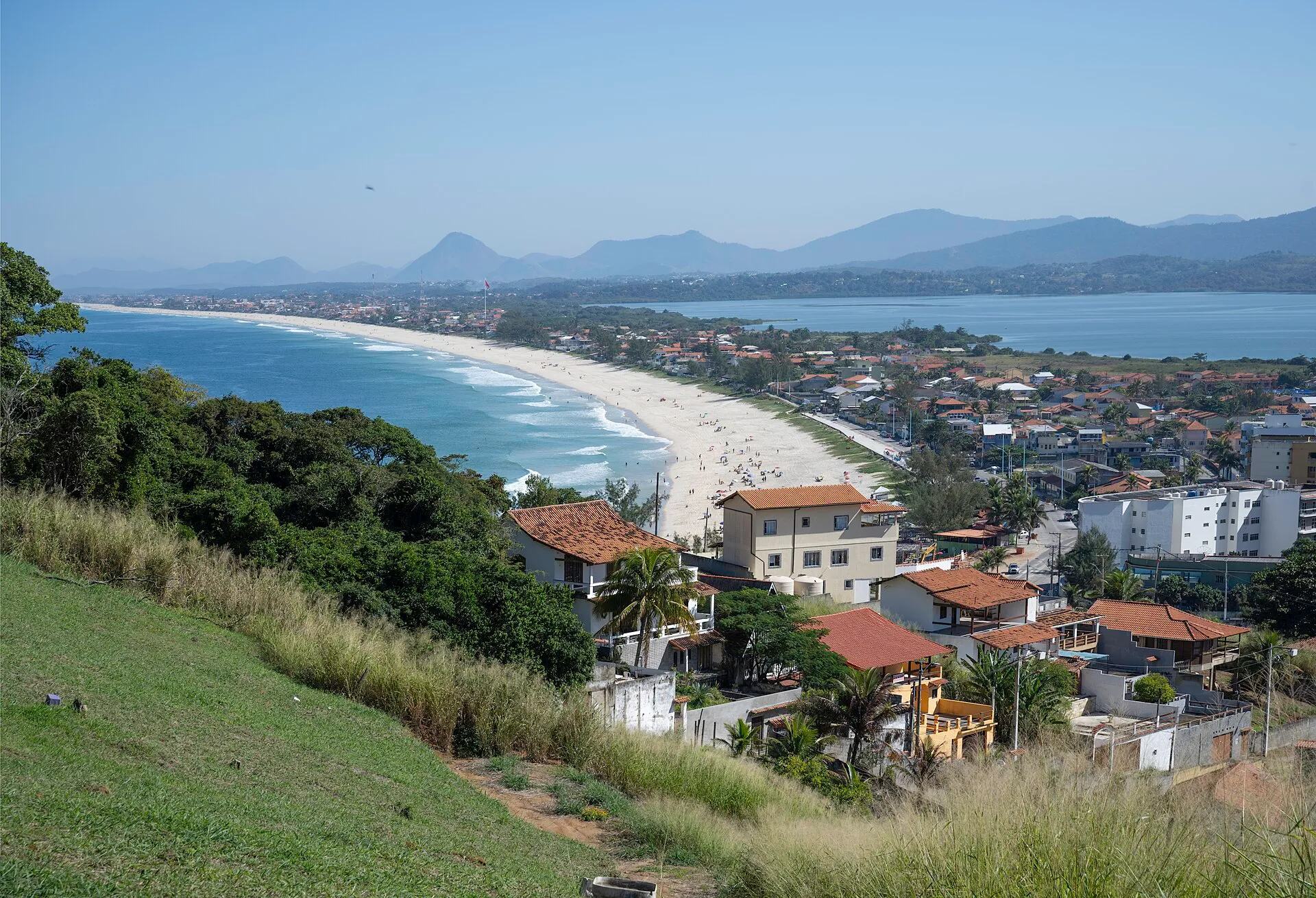 Vista panorâmica do alto do Farol de Ponta Negra, com a longa praia de areia clara, o bairro à beira-mar, a Lagoa de Guarapina e montanhas ao fundo, em Maricá