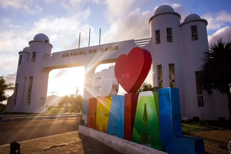 Pórtico branco de entrada de Arraial do Cabo com o letreiro colorido “Arraial” e coração vermelho ao pôr do sol