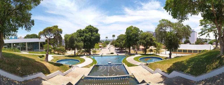 Vista panorâmica das praças Antônio Raposo e Menino João Hélio, com espelhos d’água, árvores e prédios ao redor, no centro de Araruama
