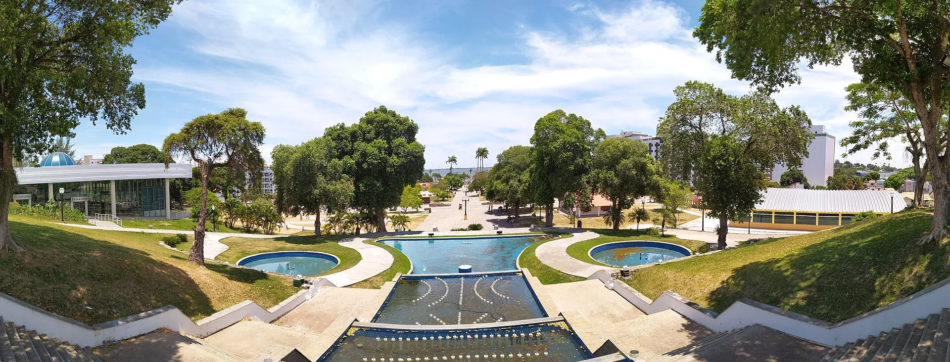 Vista panorâmica das praças Antônio Raposo e Menino João Hélio, com espelhos d’água, árvores e prédios ao redor, no centro de Araruama