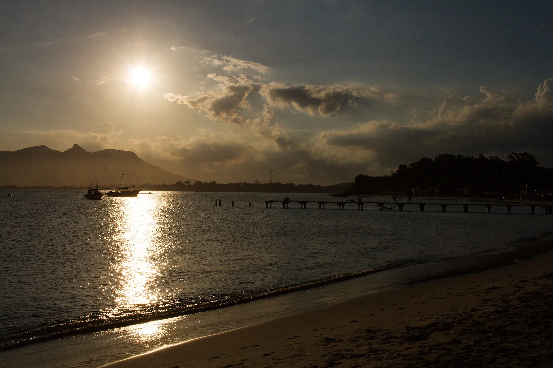 Pôr do sol sobre o mar na Praia da Boca da Barra, com barcos ancorados e um píer ao fundo